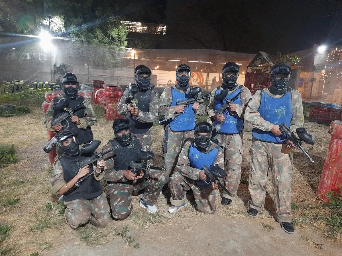 A group of young adults in camouflage gear and masks pose with paintball guns at an outdoor arena at night.