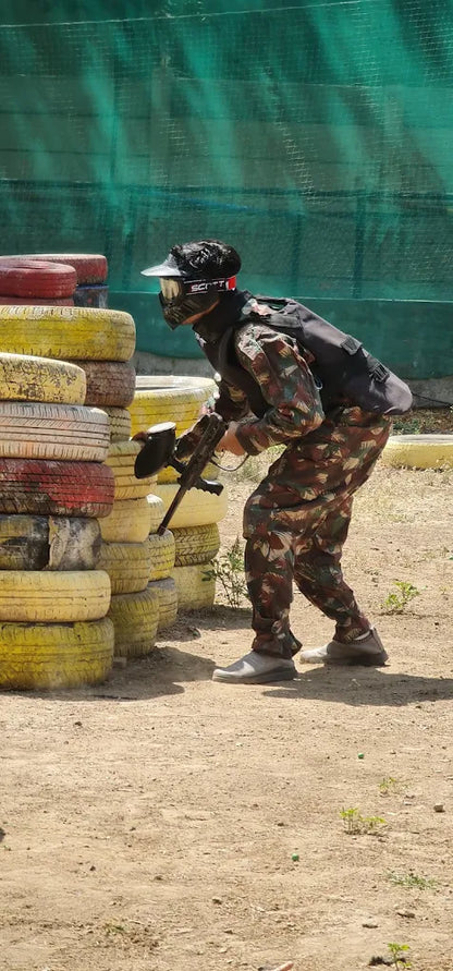 A person in camouflage gear and a mask plays paintball behind a barricade of tires.