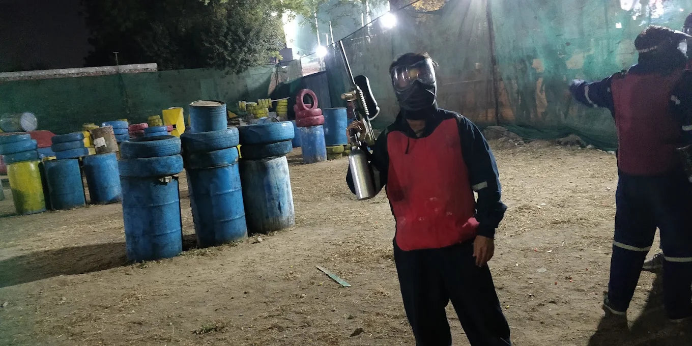 A young person in paintball gear holds a marker in an outdoor paintball arena with barrels and tires as obstacles.