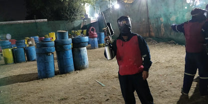 A young person in paintball gear holds a marker in an outdoor paintball arena with barrels and tires as obstacles.