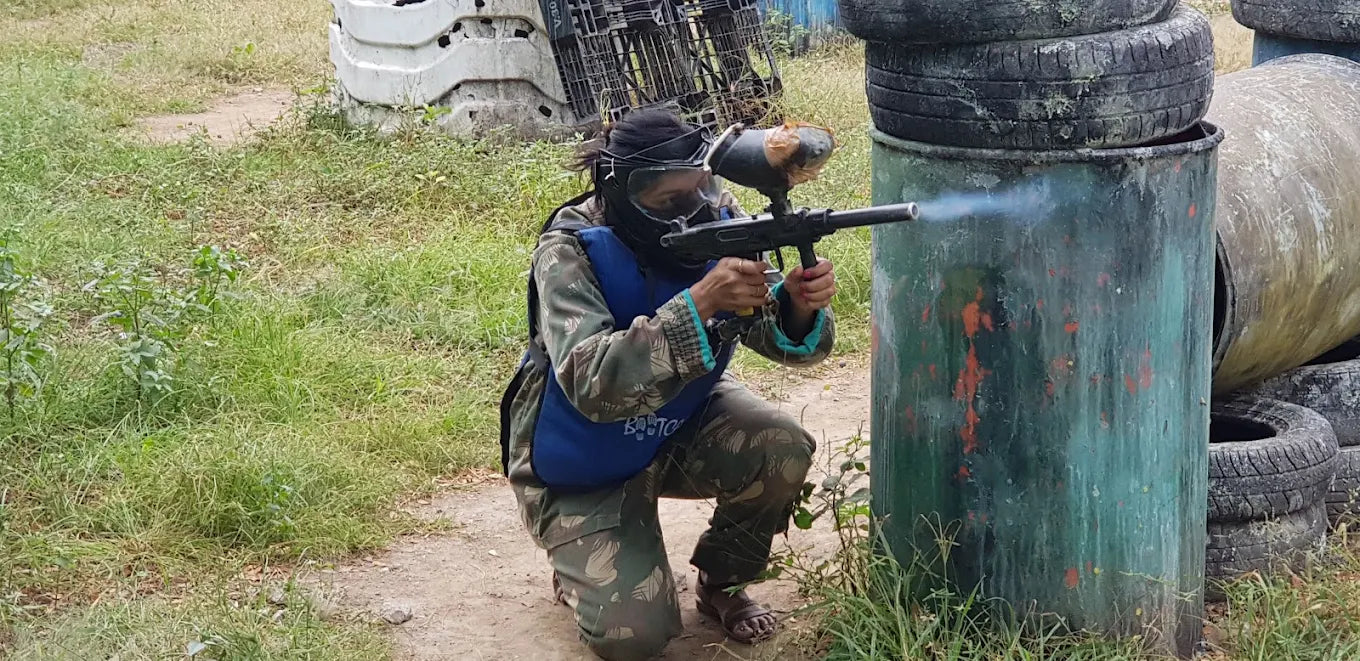 A person in camouflage and protective gear kneels and fires a paintball gun, with smoke emerging from the barrel. The scene is an outdoor paintball field with barrels and tires as obstacles.