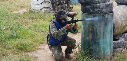 A person in camouflage and protective gear kneels and fires a paintball gun, with smoke emerging from the barrel. The scene is an outdoor paintball field with barrels and tires as obstacles.