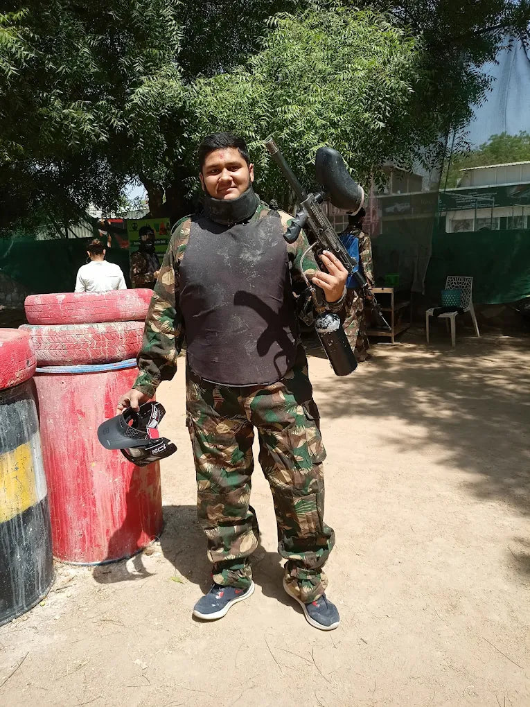 A young man in camouflage gear and a protective vest holds a paintball gun, ready for a game. He stands outdoors with trees and barriers in the background.