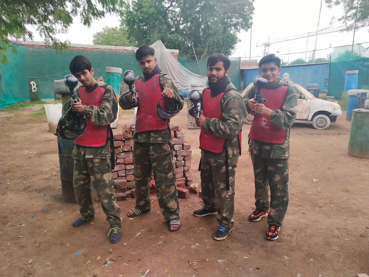 Four young men in camouflage gear and red vests stand ready for a paintball game, holding their markers.