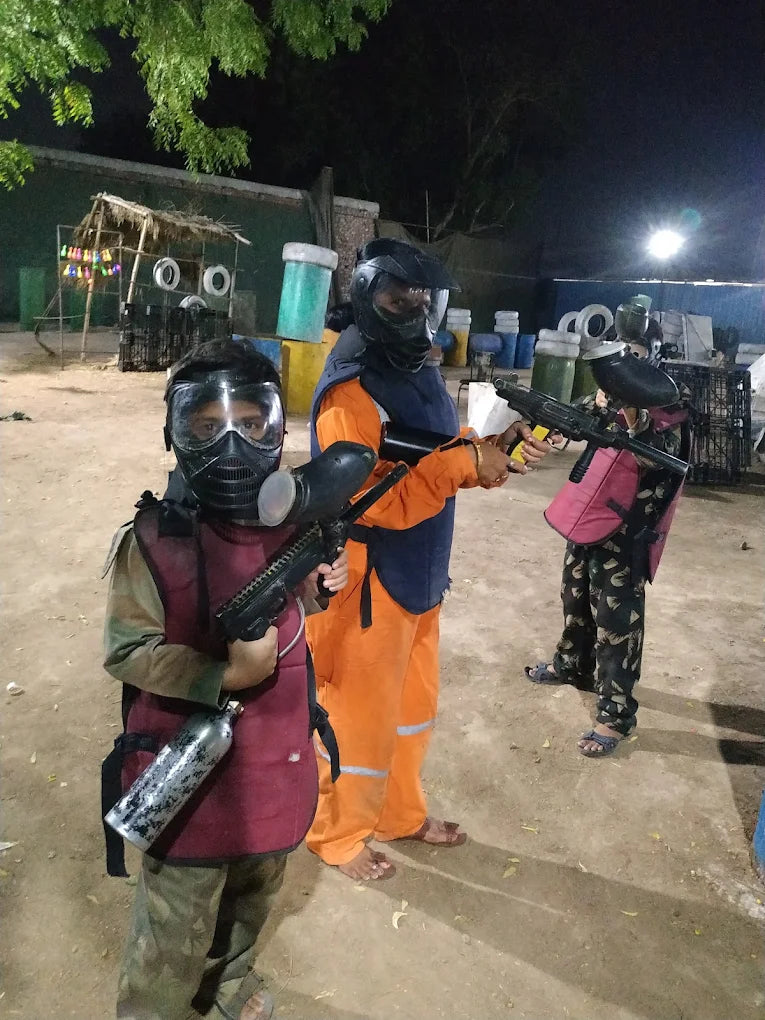 Children in paintball gear with guns stand on a dirt field at night.