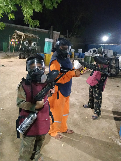 Children in paintball gear with guns stand on a dirt field at night.