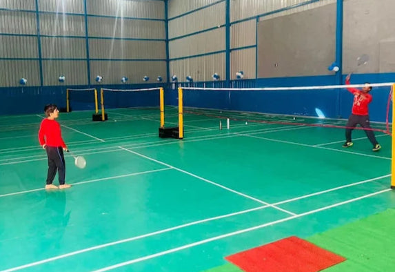 Two people playing badminton on an indoor court with green flooring and blue walls.