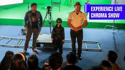 A child in a black costume stands with two adults on a blue carpet in front of a green screen.