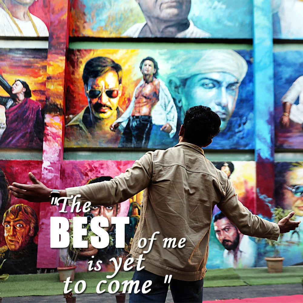 A person stands with arms outstretched before a mural of painted portraits. Text reads: "The BEST of me is yet to come".