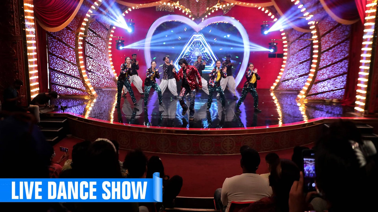 A group of young dancers performs on a brightly lit stage in front of a heart-shaped backdrop, while an audience watches from below.
