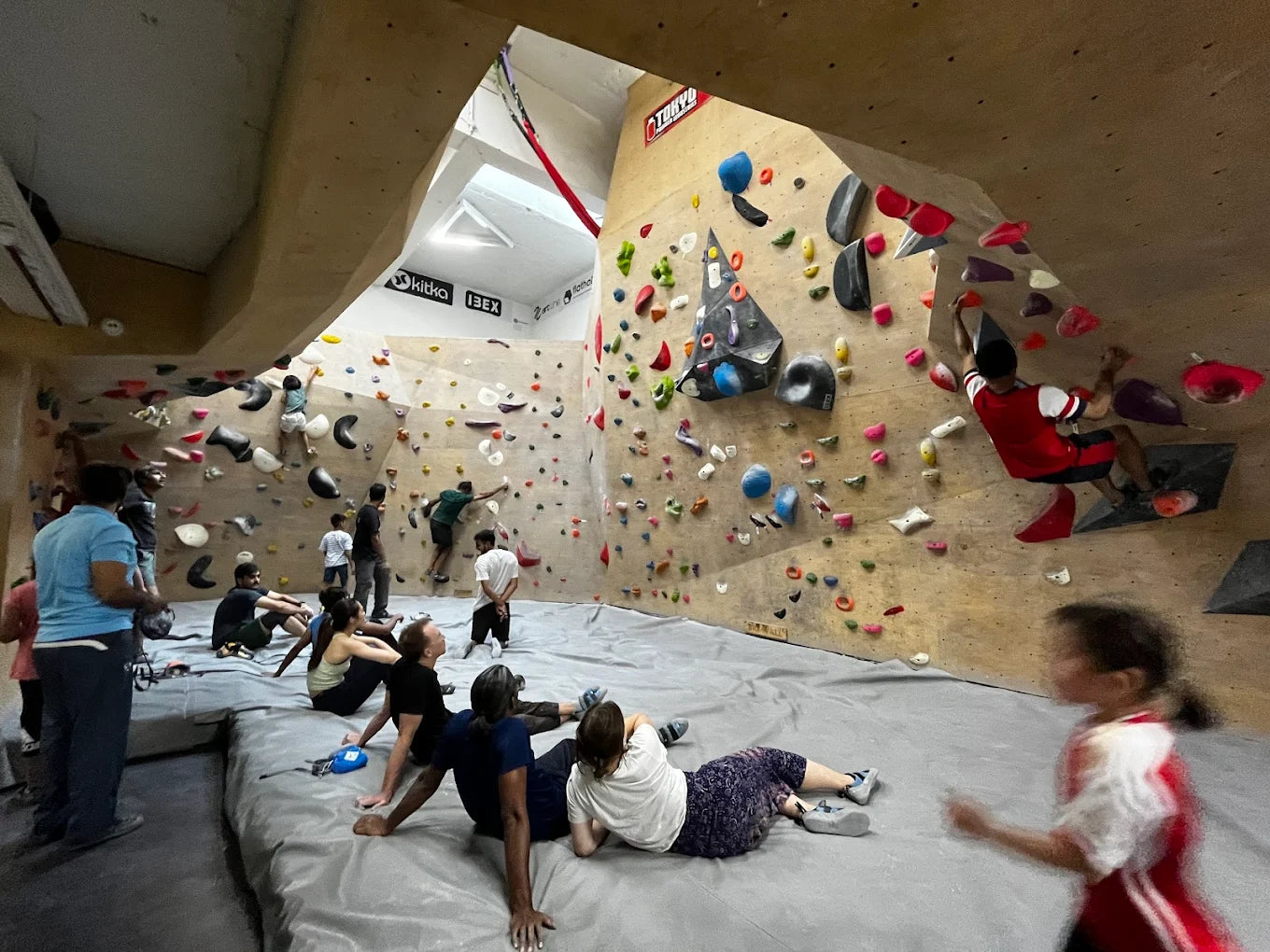 Young people boulder on a climbing wall in an indoor gym, with others resting on mats below.