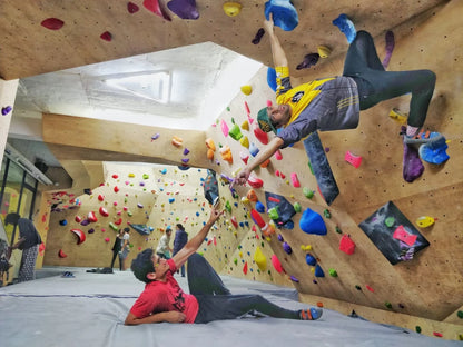 Two young men boulder in an indoor climbing gym, one hanging from the wall and the other lying on the mat.
