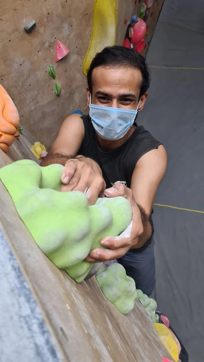 A young man in a mask climbs a bouldering wall, gripping a large green hold.