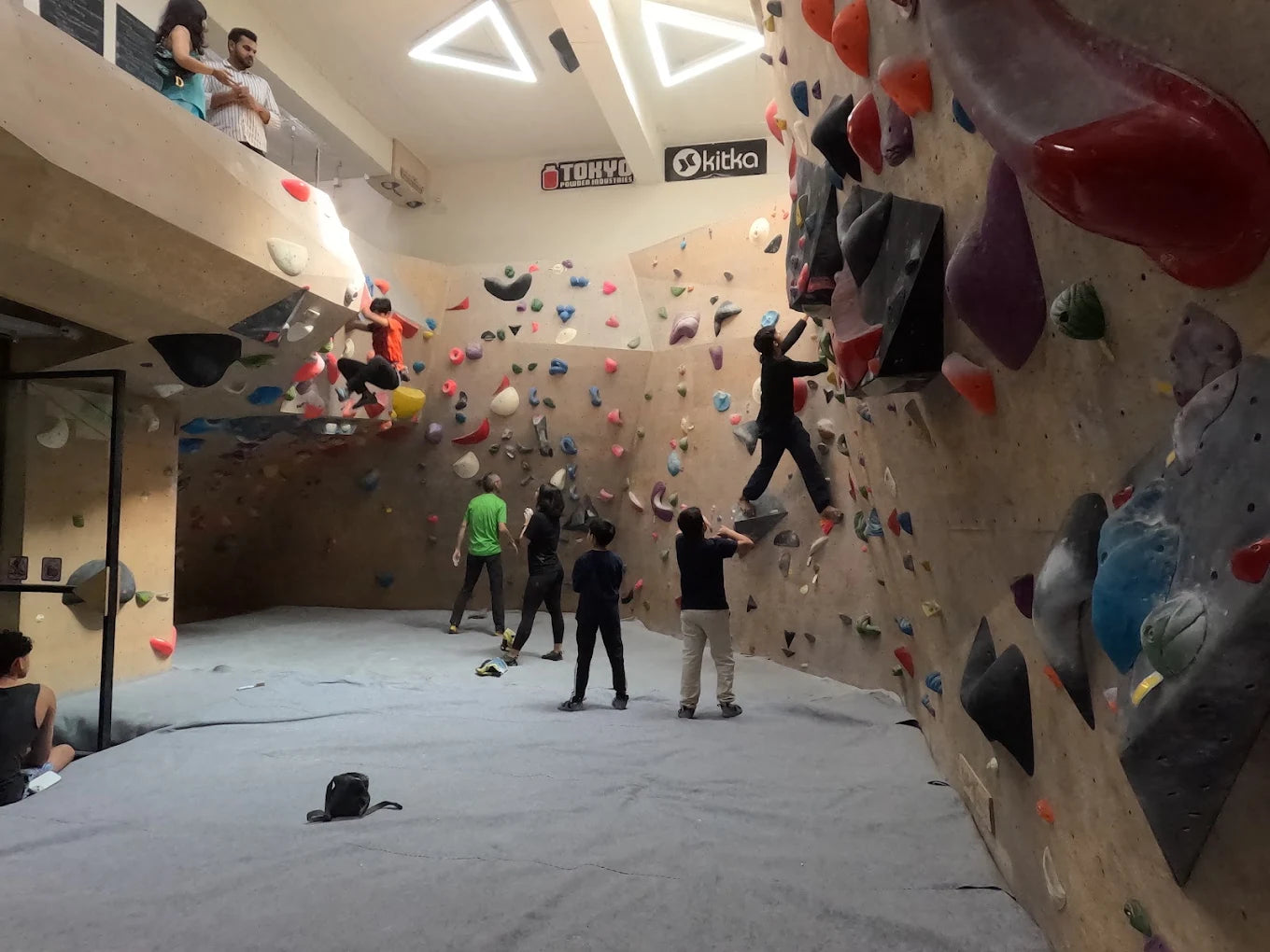 Young adults boulder at an indoor climbing gym, with some climbing the wall and others watching.