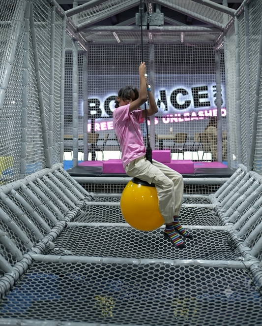 A child swings on a yellow ball in a colorful indoor playground. The child wears a pink shirt and striped socks.