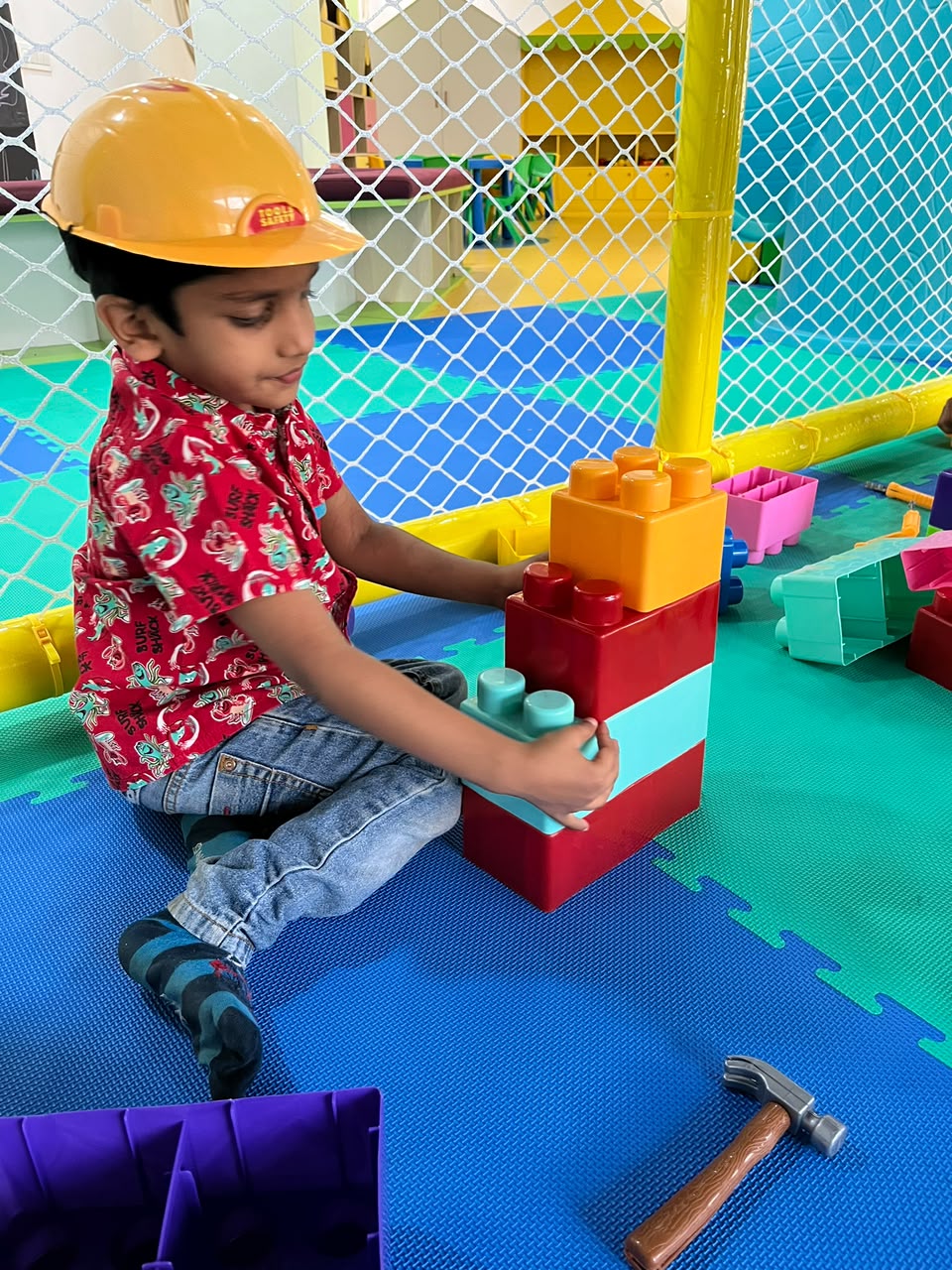 A young boy in a hard hat plays with large building blocks in a colorful indoor play area.