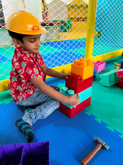 A young boy in a hard hat plays with large building blocks in a colorful indoor play area.