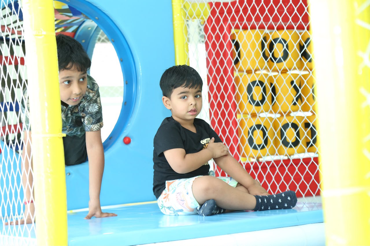 Two young boys play in a colorful indoor playground. One boy peeks from behind a blue tube, while the other sits on a blue platform.