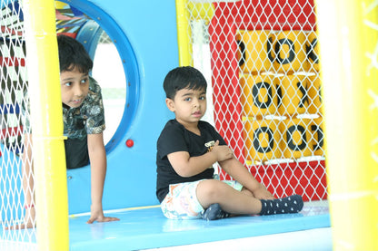 Two young boys play in a colorful indoor playground. One boy peeks from behind a blue tube, while the other sits on a blue platform.