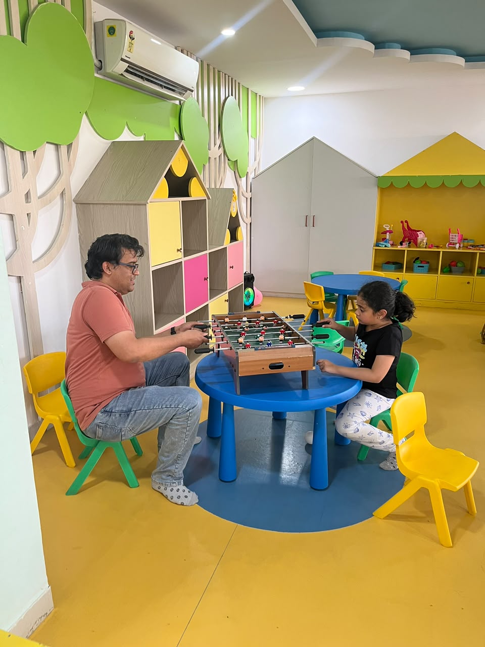 A man and a child play foosball at a colorful table in a bright, playful room.