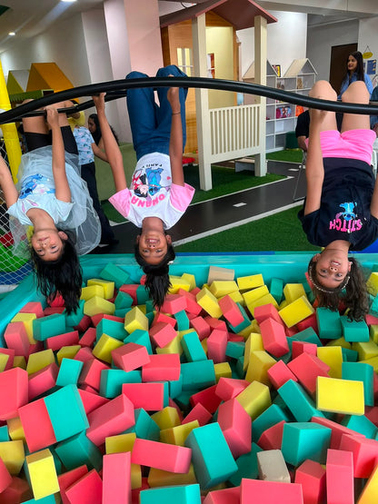 Three young girls hang upside down from a bar over a foam pit, smiling at the camera.