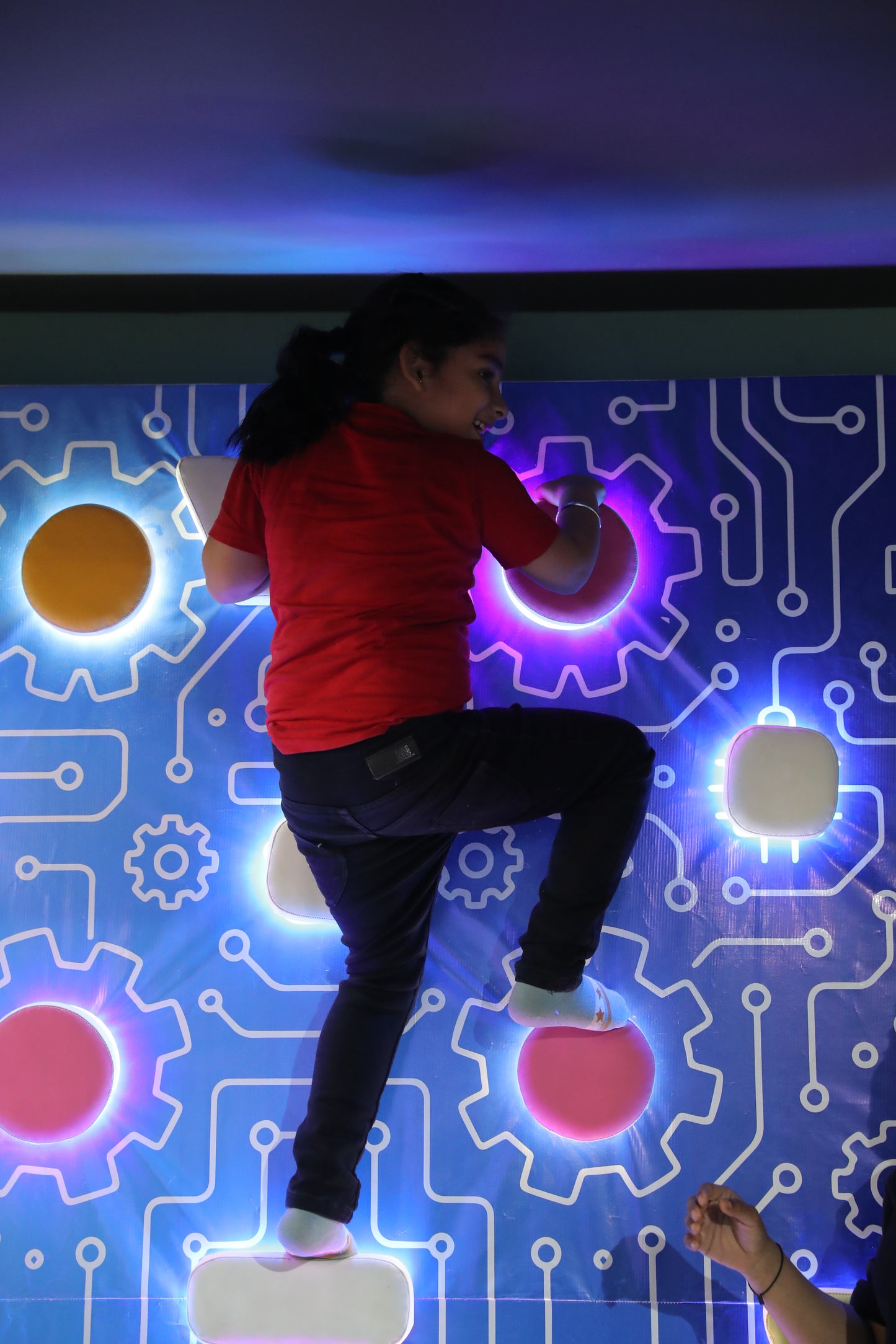 A young girl climbs a colorful, illuminated wall with a circuit board design.