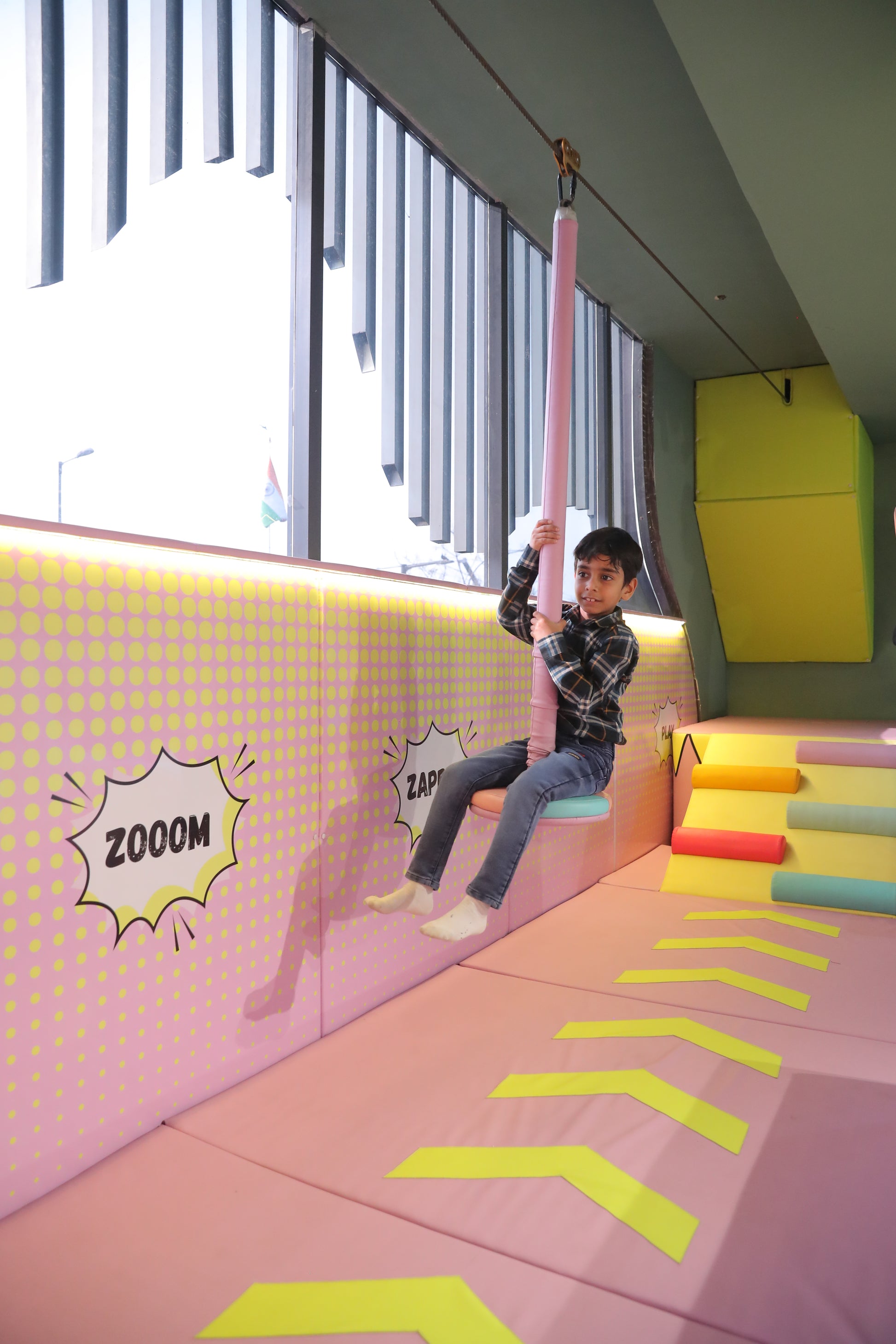 A young boy rides a zip line in a colorful indoor play area. He is smiling as he holds onto a pink padded bar.