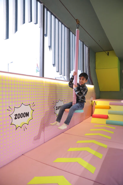 A young boy rides a zip line in a colorful indoor play area. He is smiling as he holds onto a pink padded bar.
