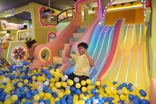 A young boy waves from a colorful ball pit slide in an indoor playground. The playground features bright lights and a large ball pit.