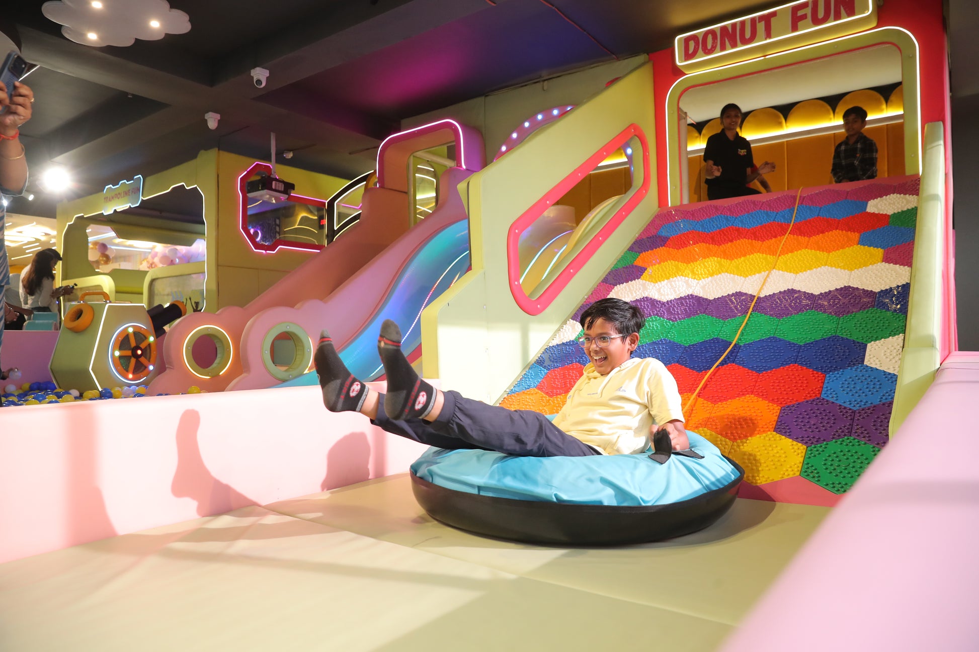A young boy slides down a colorful play structure in an indoor amusement park. He is laughing and wearing a yellow hoodie.