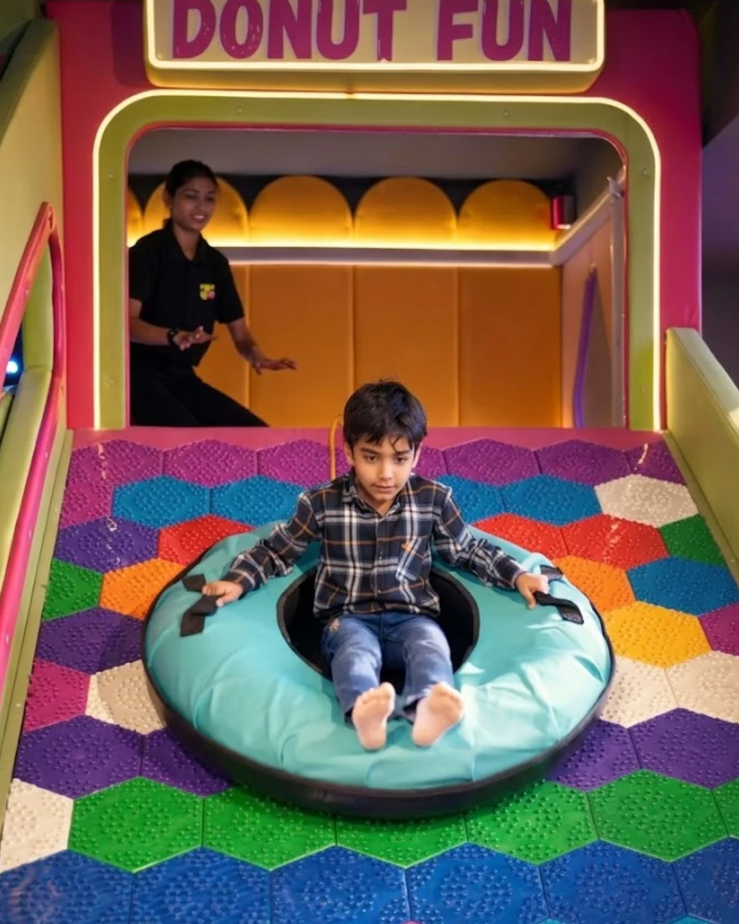A young boy slides down a colorful play area in a tube, while a woman supervises.