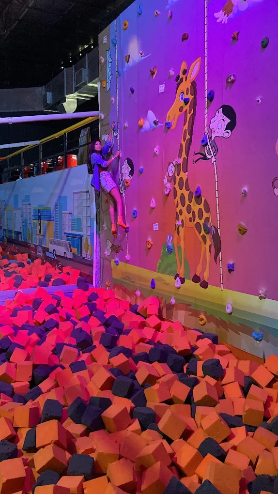 A young woman climbs a colorful rock wall above a pit of foam cubes. The wall is decorated with cartoon giraffes and characters.
