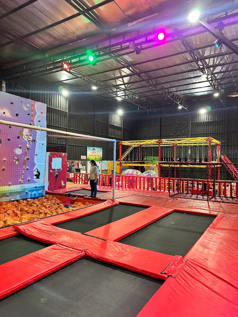 An indoor trampoline park with multiple trampolines, a climbing wall, and a foam pit. Two adults and one child are visible.