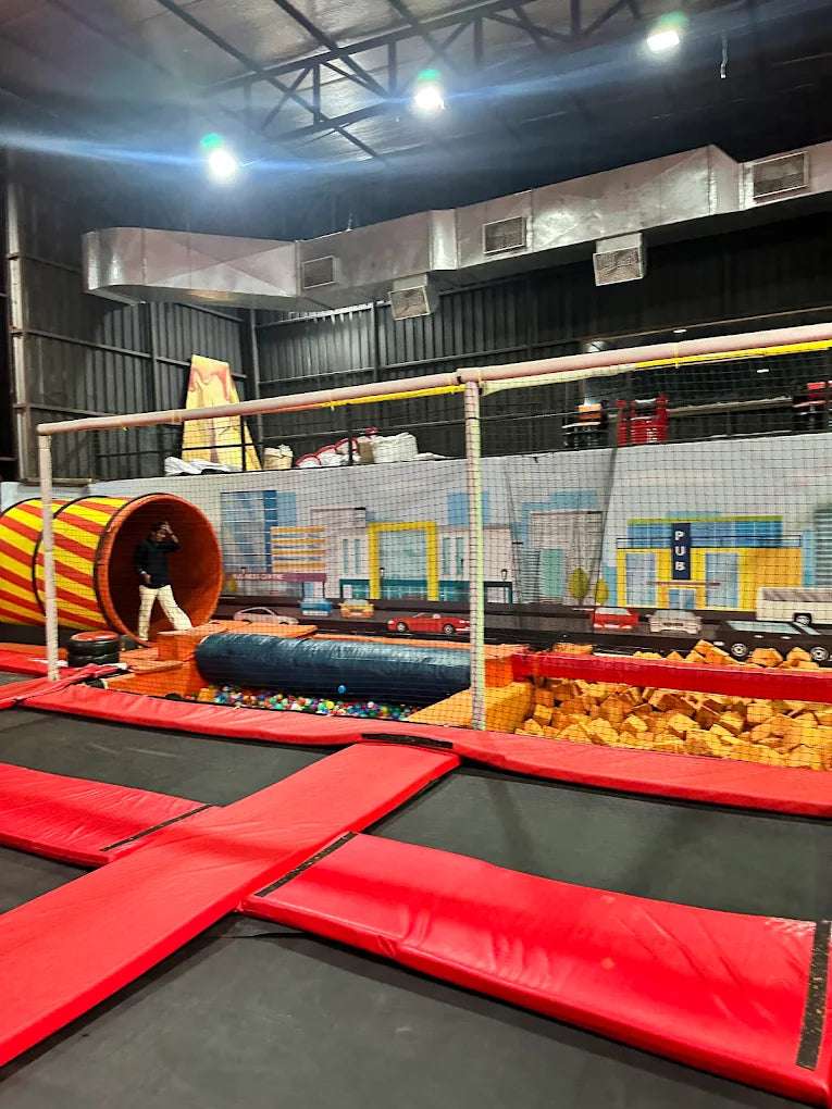 A trampoline park with red trampolines, a ball pit, and foam cubes. A child is visible near a tunnel.