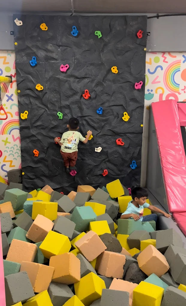 Two young boys are at a climbing wall and foam pit in an indoor play area.