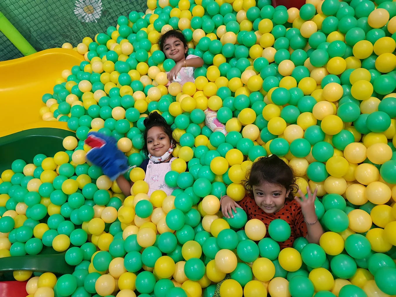Three young girls play in a ball pit filled with green and yellow balls.