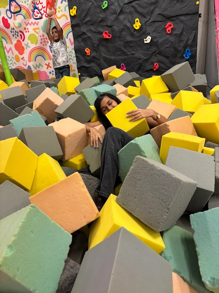 A young woman relaxes in a foam pit while a child plays on a climbing wall in the background.