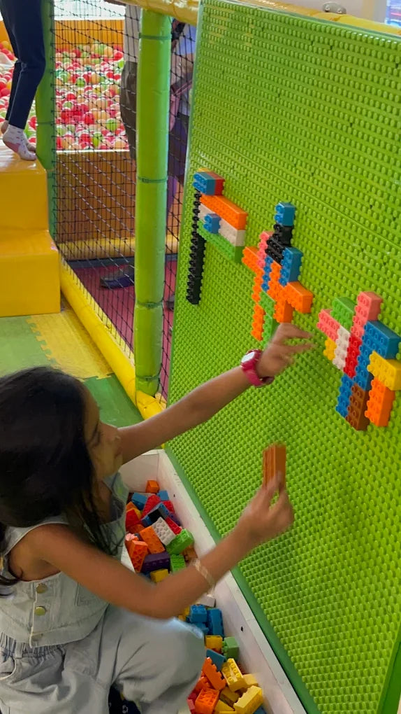 A young girl plays with colorful building blocks on a green pegboard wall in a play area.