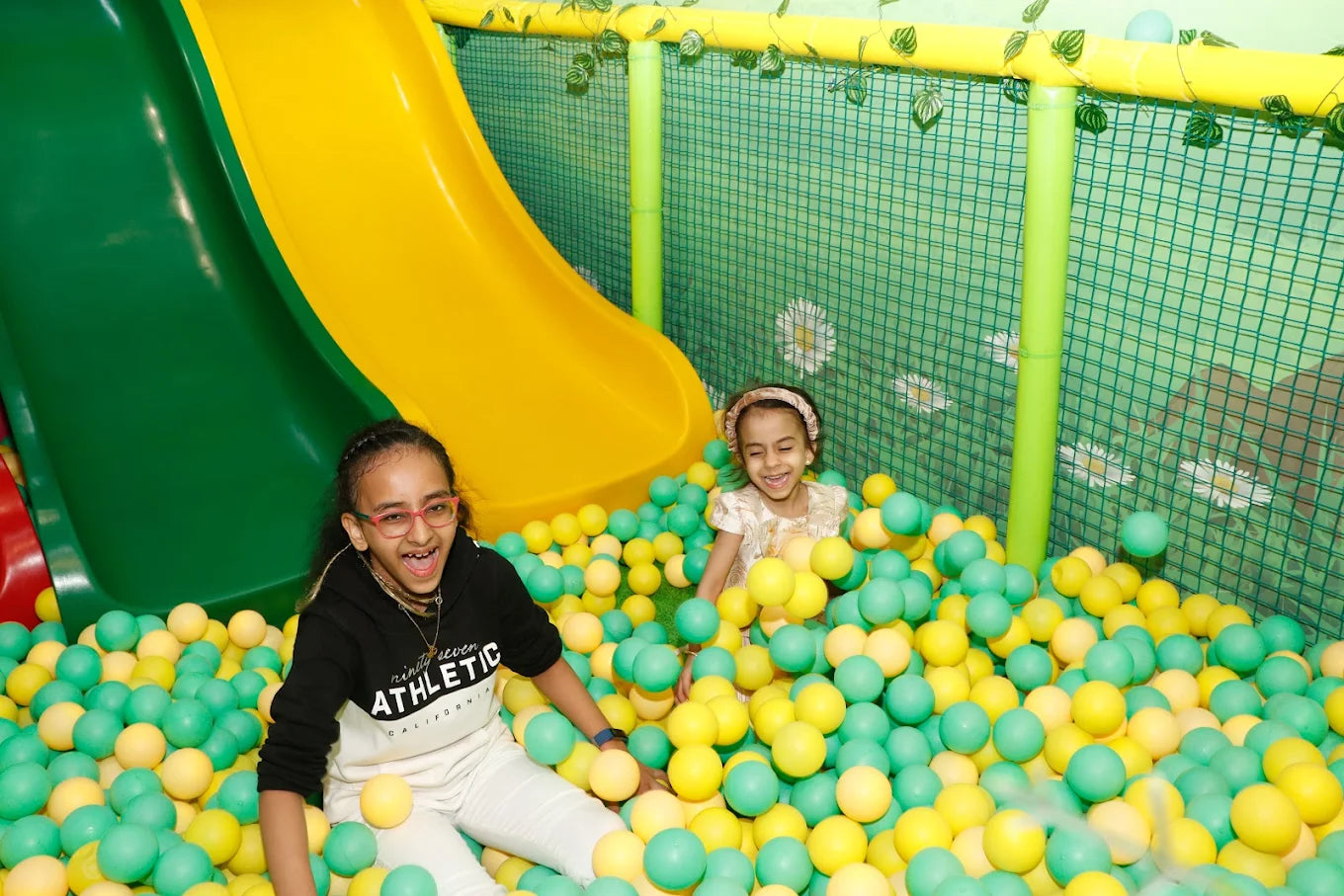 Two young girls laugh in a ball pit with a yellow and green slide.