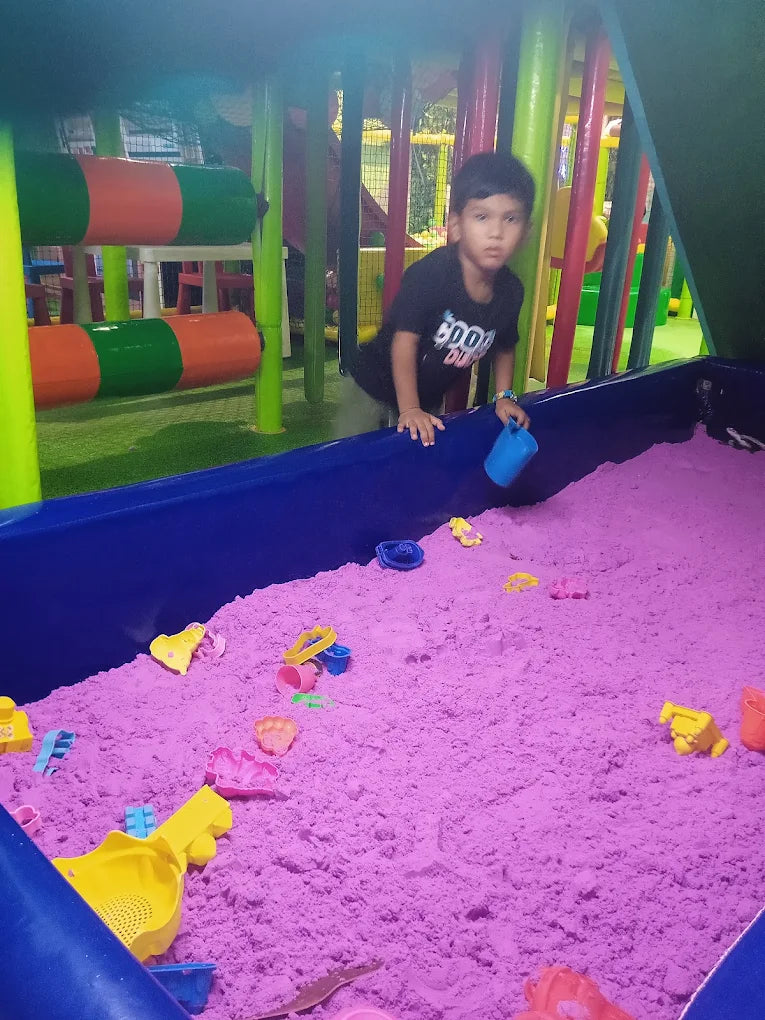 A young boy plays in a purple sandpit with toys in a colorful indoor playground.