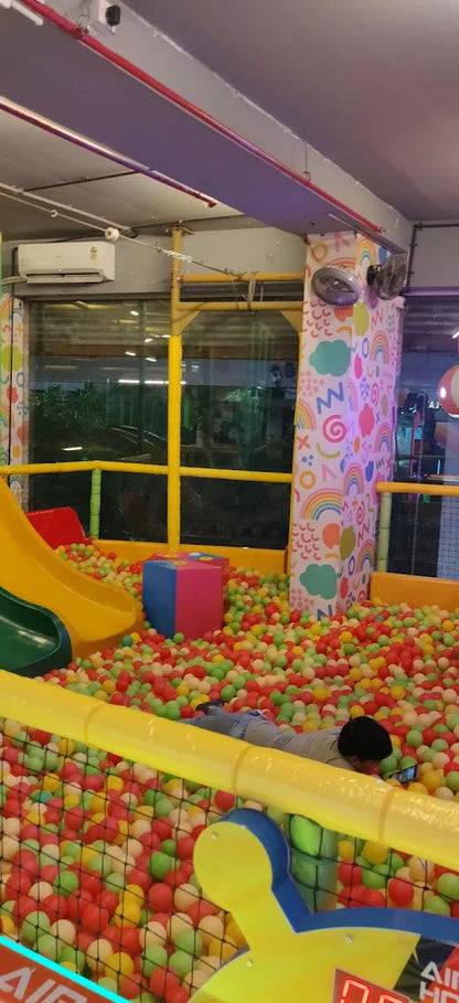 A child lies in a ball pit, playing with a phone, with a slide and colorful decorations in the background.
