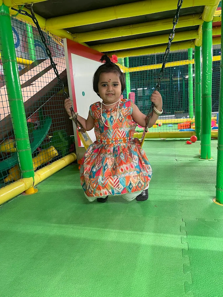 A young child in a colorful dress sits on a swing in a brightly colored indoor playground.
