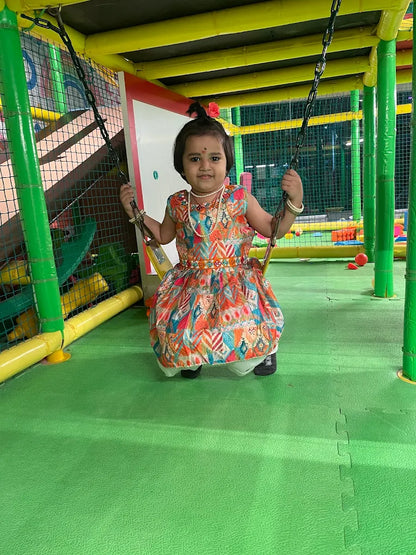 A young child in a colorful dress sits on a swing in a brightly colored indoor playground.