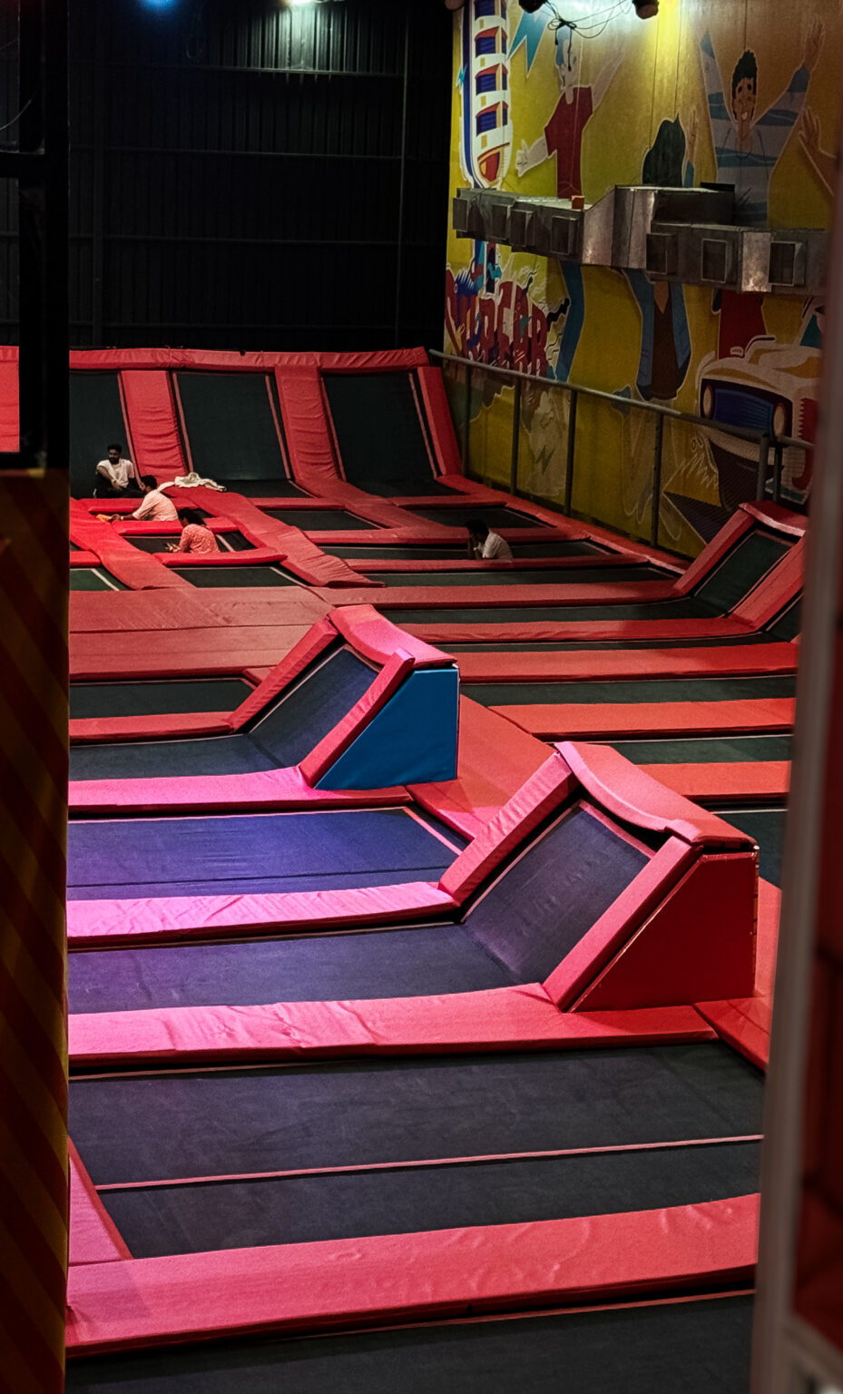 An indoor trampoline park with multiple interconnected trampolines and a few young people enjoying the space.