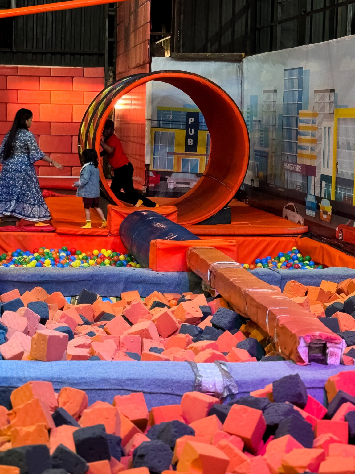 Children and adults play in a colorful indoor trampoline park with a large orange wheel and foam pit.