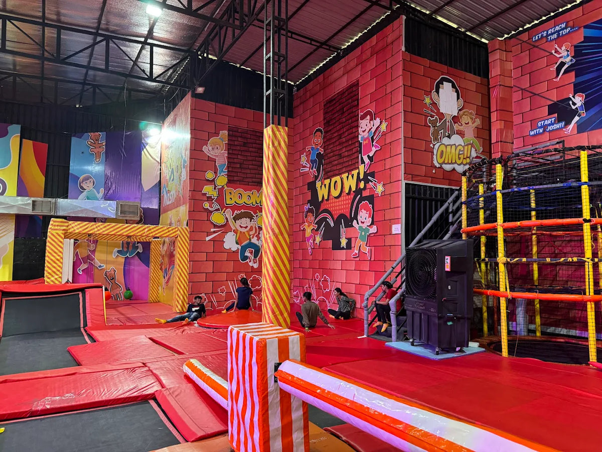 Children play in a vibrant indoor trampoline park with cartoon murals and colorful padding.