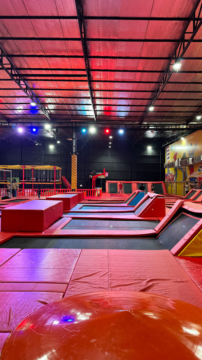 An indoor trampoline park with red padding and colorful lights, featuring children and adults jumping.
