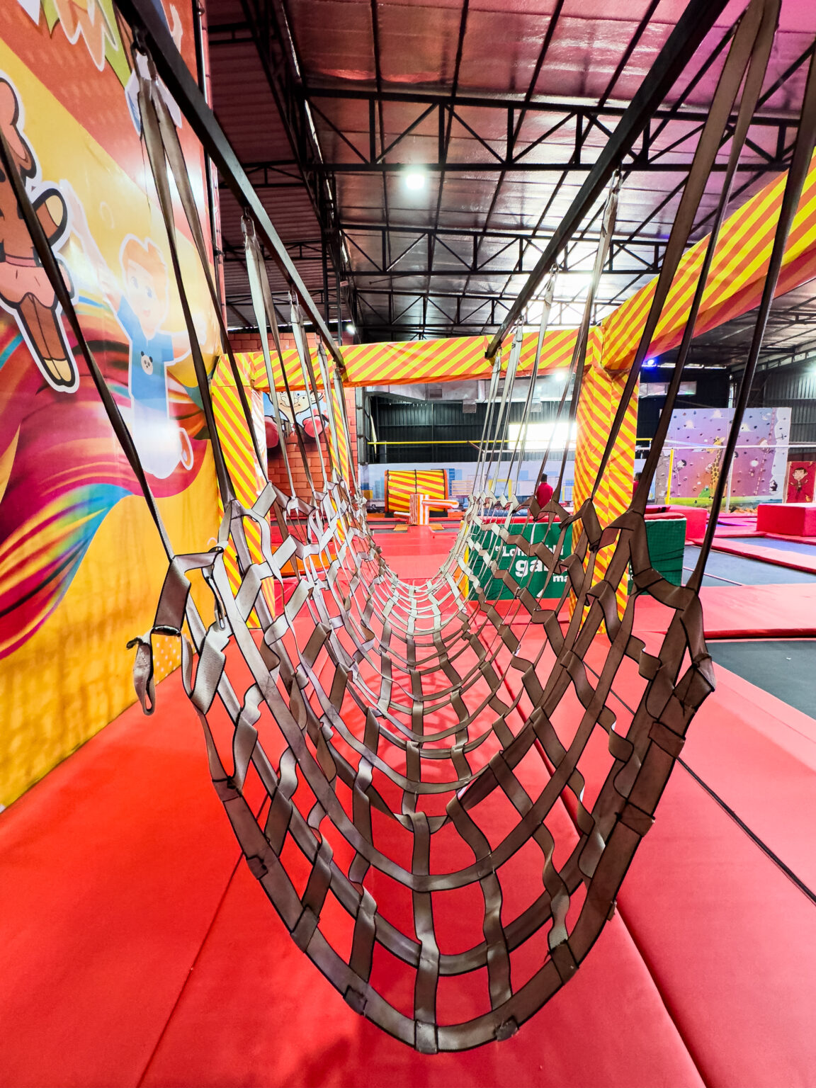 A net tunnel hangs over a trampoline park, with colorful murals and climbing walls in the background.