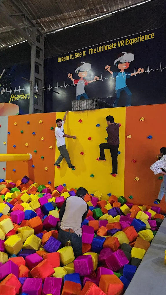 Young adults climb a colorful wall into a foam pit at an indoor adventure park.