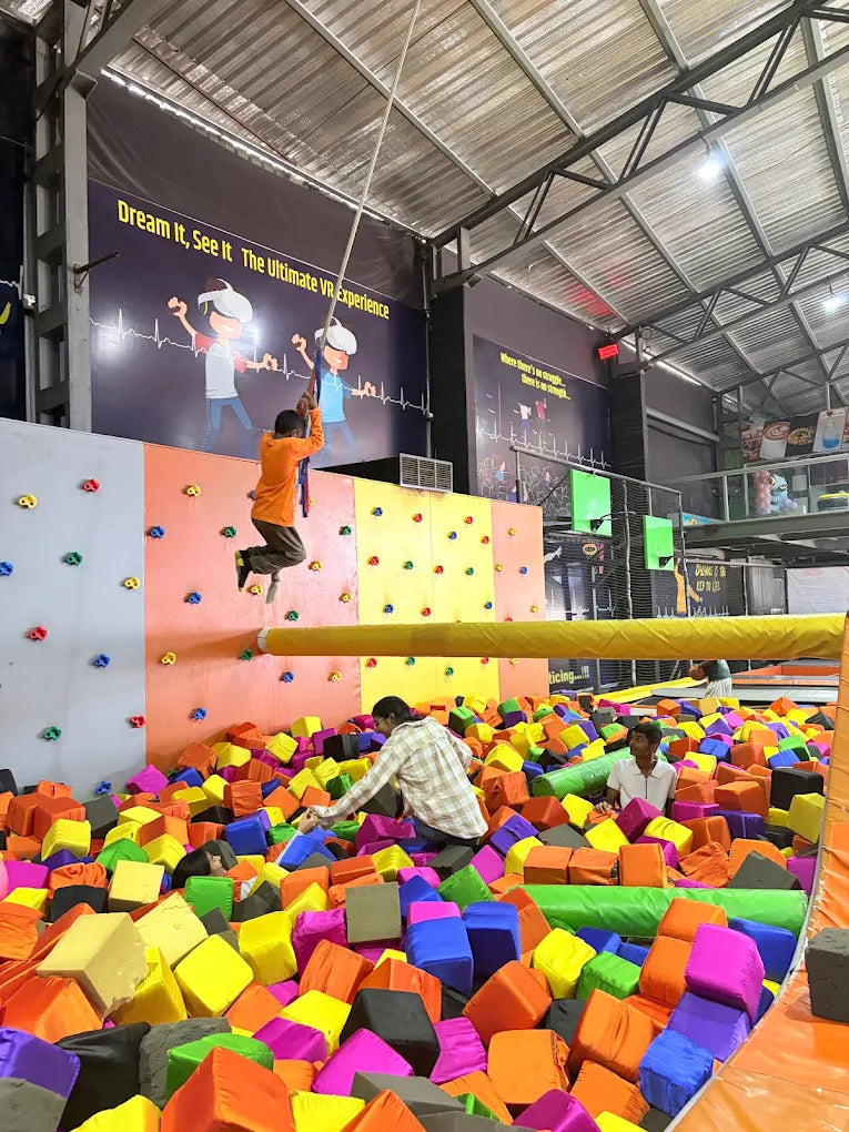 A young person swings from a rope into a foam pit at an indoor adventure park. Several people are playing in the colorful foam pit.